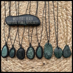 May include: A selection of dark green pebble necklaces with black cords, displayed on a woven background. A black stone sign reads "WEST COAST BEACH NATURAL PEBBLE NECKLACES". The necklaces feature various shapes and sizes of stones.