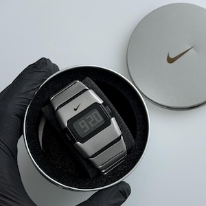 Könnte beinhalten: Eine silberfarbene Digitaluhr mit einem metallischen Gliederarmband und einem schwarzen rechteckigen Display, das die Uhrzeit 9:20 anzeigt. Die Uhr hat ein kleines Nike-Swoosh-Logo auf dem Zifferblatt und befindet sich in einem runden Metallbehälter.