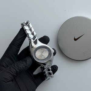 Puede incluir: Un reloj de pulsera plateado con una correa de eslabones metálicos segmentados y una esfera redonda con fondo claro y varios subesferas. Un pequeño logotipo de Nike swoosh es visible en la caja del reloj. Al lado hay una lata metálica redonda.