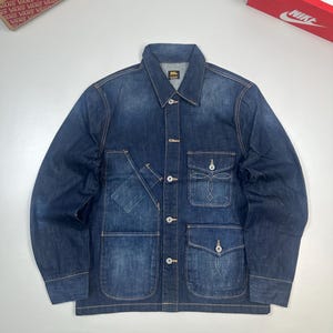 May include: A dark blue denim jacket featuring a pointed collar, long sleeves, and a button-front closure. The front includes two large lower patch pockets with flaps and a smaller chest pocket with decorative white stitching and a button closure.
