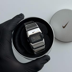 Puede incluir: Un reloj de pulsera digital plateado con correa de eslabones metálicos y una pantalla rectangular negra que muestra la hora 9:06. El reloj presenta un pequeño logotipo Nike swoosh en la esfera y se presenta en una lata redonda.