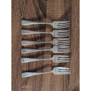 May include: Six silver forks displayed on a wooden surface. The forks have a traditional design with a rectangular handle and three tines. The forks are made of a shiny metal, likely for dining.