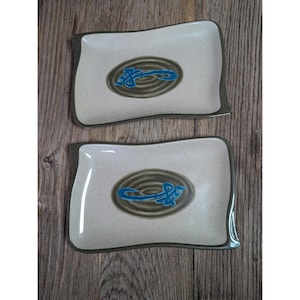 May include: Two rectangular ceramic plates with a wavy edge. The plates have a speckled off-white surface with a dark brown border. Each plate features a central oval design with a blue abstract fish symbol on a brown background.