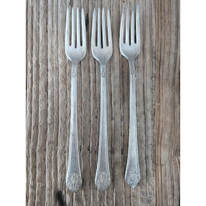 Puede incluir: Tres tenedores plateados con diseños florales ornamentados en los mangos, colocados sobre una superficie de madera envejecida. Los tenedores tienen púas y mangos largos y delgados. La estética general es vintage y rústica.