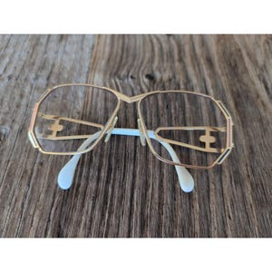 May include: Gold-tone eyeglasses with a geometric frame design. The glasses have clear lenses, white nose pads, and a gold bridge. The temples are also gold with a decorative element. The frame has accents of black and pink.