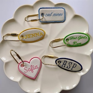 Könnte beinhalten: Eine Sammlung von bestickten Schlüsselanhängern in verschiedenen Formen, darunter ein Rechteck, ein Oval und ein Herz. Jeder Schlüsselanhänger zeigt Text wie "cat mom", "Jenna", "Hot Girls Run", "Sofia" und "ASP". Die Schlüsselanhänger haben goldfarbene Hardware.