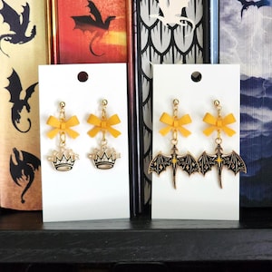 May include: Two pairs of earrings displayed on white cards. One pair features yellow bow accents and crown charms. The other pair has yellow bows and black dragon wing charms. Both pairs have gold-colored hardware.