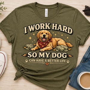Puede incluir: Una camiseta de cuello redondo color verde oliva con el gráfico de un golden retriever con pañuelo rojo, rodeado de fajos de billetes. El texto dice: I WORK HARD SO MY DOG CAN HAVE A BETTER LIFE. El diseño incluye estrellas y huellas.