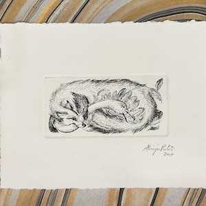 Puede incluir: Un grabado en blanco y negro de un animal durmiendo acurrucado alrededor de un pájaro. La obra de arte está sobre un trozo de papel rectangular con un borde rugoso. La firma del artista y el año 2017 son visibles.