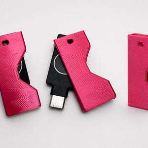 May include: A collection of pink USB drive covers with a unique, angular design. One cover is paired with a black USB drive. Each cover has a small heart-shaped hole at the top. The items are arranged on a white surface.