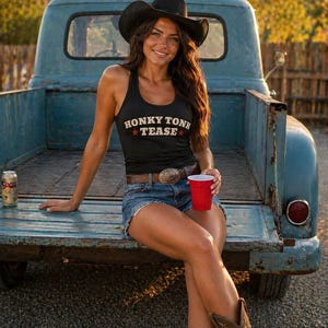 Puede incluir: Una persona con una camiseta sin mangas negra con las palabras "HONKY TONK TEASE" en blanco y rojo, pantalones cortos de mezclilla, un sombrero de vaquero y botas, sentada en la puerta trasera de una camioneta azul vintage. Se ve una taza roja y una lata de cerveza.