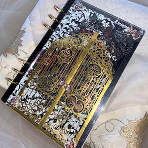 May include: A disc-bound notebook featuring a black and white floral cover with a raven and skull illustration. A gold-toned, intricate, laser-cut metal overlay shaped like a gate sits on top, accented with small, multi-colored iridescent crystals.