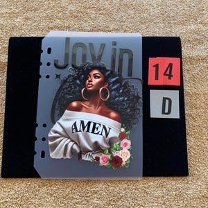 Peut inclure: Une couverture de planificateur translucide présentant une illustration graphique d'une personne aux cheveux noirs bouclés, boucles d'oreilles créoles dorées et un haut blanc à épaules dénudées avec le texte AMEN. Le texte Joy in est visible.