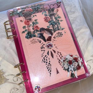 May include: A pink acrylic binder featuring gold-tone metal rings and a decorative cover insert. The insert displays a couple standing between stone columns surrounded by roses, a large laser-cut bow, a floral bouquet, and iridescent rhinestones.