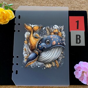 May include: A translucent plastic planner divider featuring a colorful illustration of a whale with blue and yellow patterns, surrounded by floral motifs. The divider has a vertical row of punched holes on the left side and the text 1 B on the right.