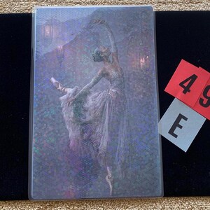 May include: A rectangular holographic trading card featuring a ballerina in a flowing dress performing a dance pose. The image has a shimmering, multi-colored dot pattern overlay. Two small red and grey paper tags with the characters 49 and E are placed next to the card.