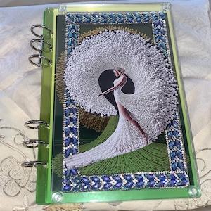 May include: A rectangular binder with a clear cover featuring a decorative border of blue and silver rhinestones. The center image depicts a figure in a white gown with a large, circular, textured white train against a green landscape.