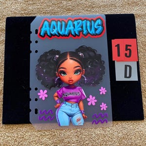 May include: A translucent planner dashboard featuring a cartoon illustration of a character with dark curly hair in pigtails, wearing a purple shirt with the text AQUARIUS and light blue jeans. The word AQUARIUS appears at the top in blue.