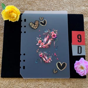 Könnte beinhalten: Ein durchscheinendes, rechteckiges Planer-Dashboard mit einem Aufdruck von zwei hochhackigen Schuhen, die mit rosa und roten Blumen verziert sind, umgeben von mehreren schwarzen und leopardenmuster-Herzsymbolen. Die linke Kante hat sechs Stanzlöcher.