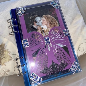 May include: A blue acrylic ring-bound notebook featuring a cover illustration of a masked figure and a woman. The cover is decorated with a large purple die-cut bow, intricate black filigree accents, and blue rhinestone embellishments at corners.