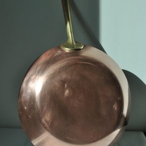 May include: A copper frying pan with a brass handle. The pan is round with a shiny, reflective surface. The handle is attached with rivets. The pan is designed for cooking.