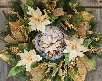 May include: A circular wreath featuring green pine branches, gold glittered ribbons, and gold berry clusters. Three large cream-colored poinsettia flowers are arranged around a central circular plaque decorated with a painted poinsettia design.
