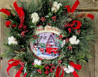 May include: A Christmas wreath featuring green pine needles, holly leaves, red berries, and white cotton bolls. A central circular sign displays a snowy scene with a red pickup truck, a snowman, and the text Merry Christmas in red lettering.