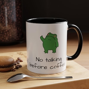 May include: White ceramic coffee mug with a black interior and handle. The mug features a green cartoon frog illustration and the text "No talking before coffee" in black. A spoon and a macaron are in the foreground.