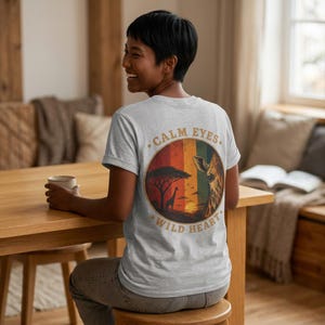 Könnte beinhalten: Hellgraues T-Shirt mit einem kreisförmigen Aufdruck auf dem Rücken, der eine Giraffe, einen Akazienbaum und einen Sonnenuntergang zeigt. Der Text "CALM EYES WILD HEART" ist im Vintage-Stil um die Grafik gedruckt. Das Shirt hat kurze Ärmel.