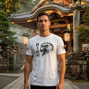 Könnte beinhalten: Weißes T-Shirt mit einem schwarz-weißen Adlerkopf-Design mit blauen Augen. Das Wort "HIGH" ist vertikal auf der linken Seite aufgedruckt. Eine kleine Grafik befindet sich auf dem linken Ärmel.
