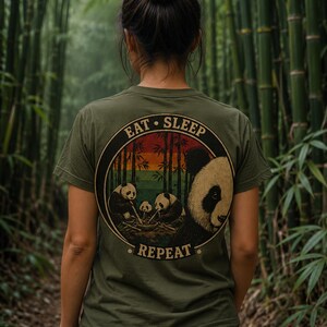 Könnte beinhalten: Olivgrünes T-Shirt mit einem kreisförmigen Aufdruck, der Pandas und Bambus zeigt. Der Aufdruck enthält den Text "EAT SLEEP REPEAT". Die Person trägt das T-Shirt in einem Bambuswald.