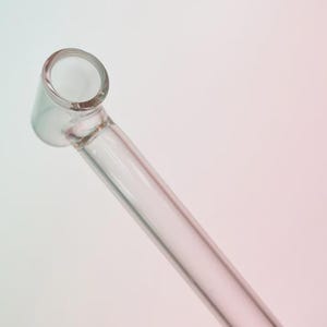 May include: A clear glass pipe with a rounded bowl and a long, slender stem. The pipe is angled, with the bowl facing upwards. The glass is transparent, allowing a view through the object. The background is a gradient of pink and white.