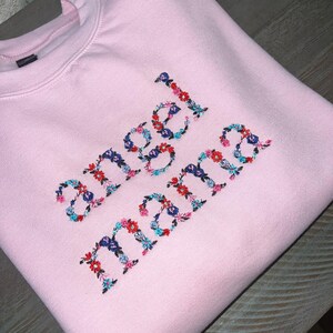 May include: A light pink sweatshirt with the words "angel mama" embroidered in colorful floral letters. The embroidery features red, blue, and teal flowers. The sweatshirt is folded and appears to be made of a soft material.