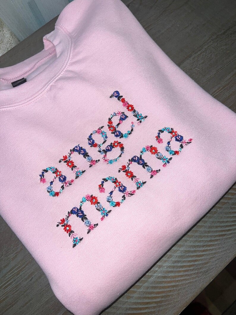 May include: A light pink sweatshirt with the words "angel mama" embroidered in colorful floral letters. The embroidery features red, blue, and teal flowers. The sweatshirt is folded and appears to be made of a soft material.