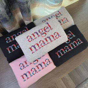 May include: A collection of folded sweatshirts in various colors, including black, gray, and pink. Each sweatshirt features the words "angel mama" embroidered in a floral font, with a mix of red, pink, purple, and blue flowers.