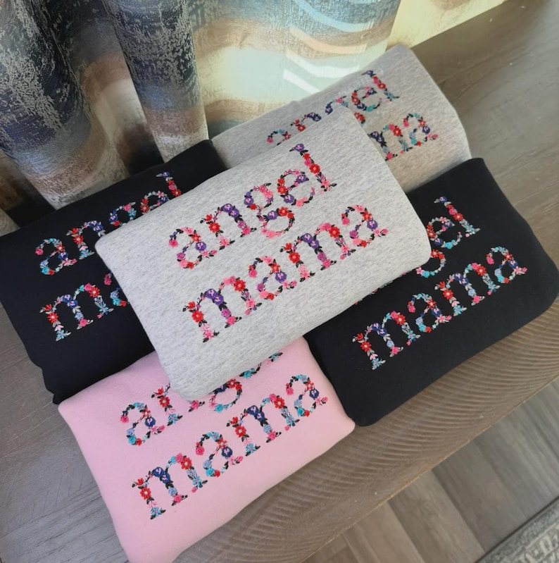 May include: A collection of folded sweatshirts in various colors, including black, gray, and pink. Each sweatshirt features the words "angel mama" embroidered in a floral font, with a mix of red, pink, purple, and blue flowers.