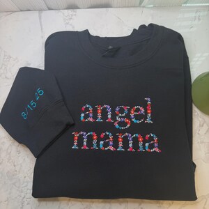 May include: Black sweatshirt with the words "angel mama" embroidered in colorful floral patterns. The left sleeve cuff has the date "8/15/25" embroidered in teal. The sweatshirt is made of a soft material.