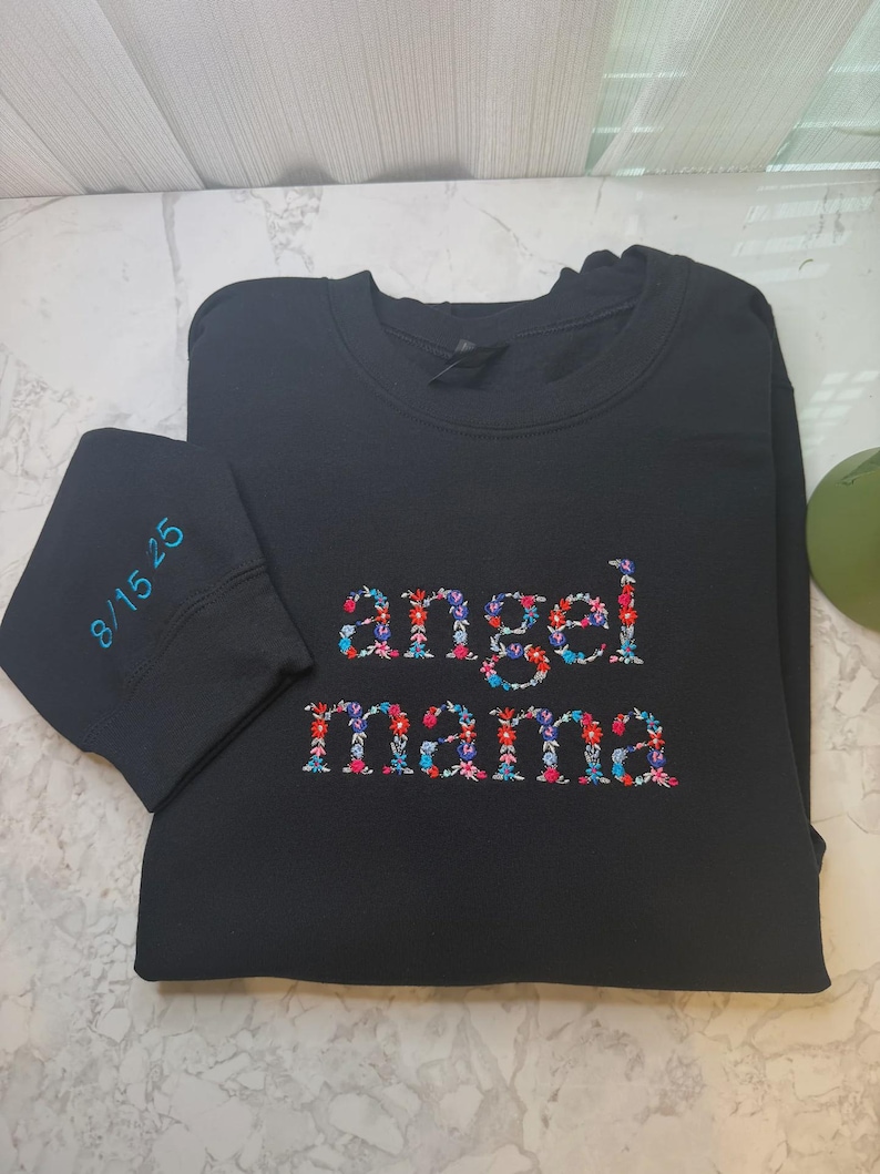 May include: Black sweatshirt with the words "angel mama" embroidered in colorful floral patterns. The left sleeve cuff has the date "8/15/25" embroidered in teal. The sweatshirt is made of a soft material.