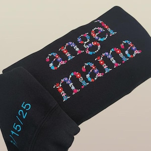 May include: Black sweatshirt with the words "angel mama" embroidered in colorful floral letters. The sleeve has the numbers "15/25" embroidered in teal. The sweatshirt is folded.