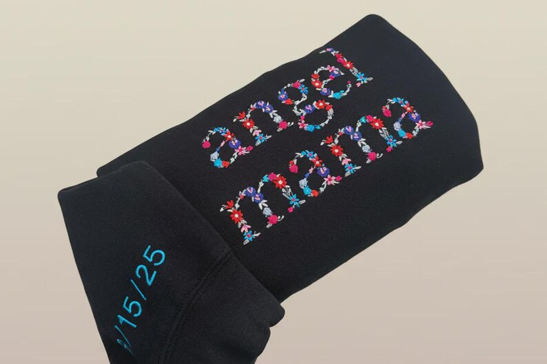 May include: Black sweatshirt with the words "angel mama" embroidered in colorful floral letters. The sleeve has the numbers "15/25" embroidered in teal. The sweatshirt is folded.