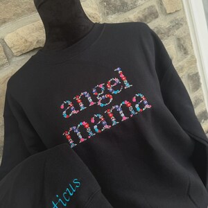 May include: Black sweatshirt with the words "angel mama" embroidered in colorful floral letters. The sleeve features the name "Atticus" embroidered in blue. The sweatshirt is displayed on a black mannequin.
