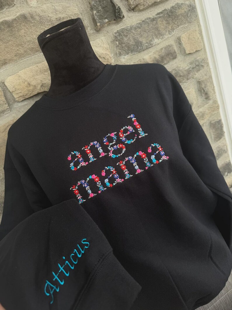 May include: Black sweatshirt with the words "angel mama" embroidered in colorful floral letters. The sleeve features the name "Atticus" embroidered in blue. The sweatshirt is displayed on a black mannequin.