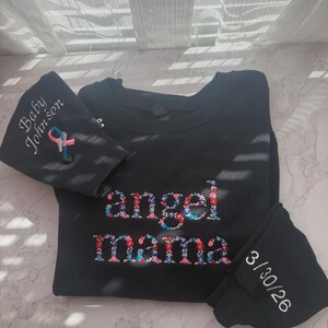 May include: Black sweatshirt with floral embroidered "angel mama" text. The left sleeve has "Baby Johnson" and a ribbon design. The right sleeve has the date "3/30/26" embroidered in white.