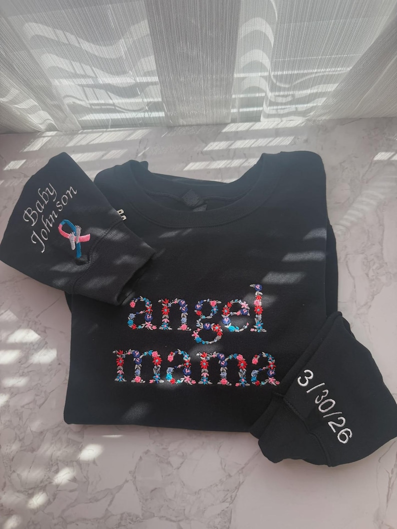 May include: Black sweatshirt with floral embroidered "angel mama" text. The left sleeve has "Baby Johnson" and a ribbon design. The right sleeve has the date "3/30/26" embroidered in white.