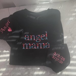 May include: Black long-sleeved shirt with the words "angel mama" embroidered in a floral design. The sleeve has the word "Angel" and a pink heart. A matching face mask is included, embroidered with dates.