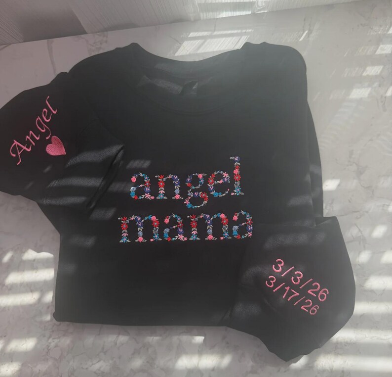 May include: Black long-sleeved shirt with the words "angel mama" embroidered in a floral design. The sleeve has the word "Angel" and a pink heart. A matching face mask is included, embroidered with dates.