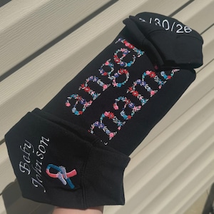 May include: Black baby beanie with floral embroidery spelling out "Baby Johnson" and "Angel Baby". A pink and blue ribbon is embroidered on the side. The top of the beanie has the numbers "-130/26" embroidered in white.