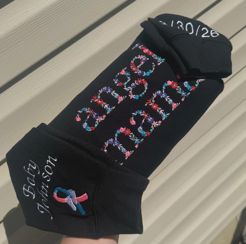 May include: Black baby beanie with floral embroidery spelling out "Baby Johnson" and "Angel Baby". A pink and blue ribbon is embroidered on the side. The top of the beanie has the numbers "-130/26" embroidered in white.