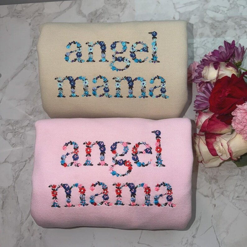 May include: Two embroidered sweatshirts. The top one is beige, the bottom one is pink. Both have the words "angel mama" embroidered in a floral design with blue, red, and white flowers.
