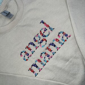 May include: A beige sweatshirt with the embroidered words "angel mama" in colorful floral lettering. The embroidery features a mix of red, blue, and pink flowers, creating a vibrant contrast against the neutral background. The sweatshirt has a crew neck and long sleeves.